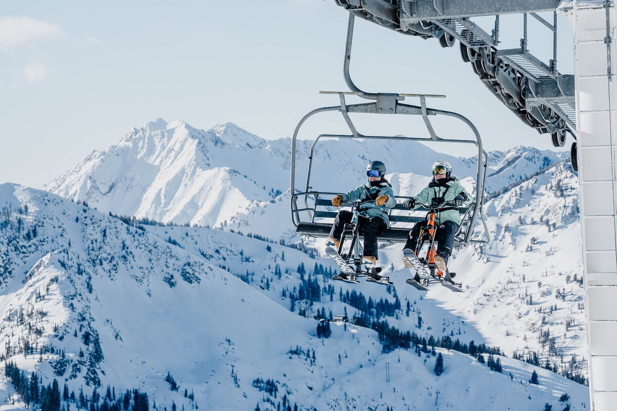 Riders loading and riding a three-ski ski bike onto a chairlift, frame hook resting on the chair seat