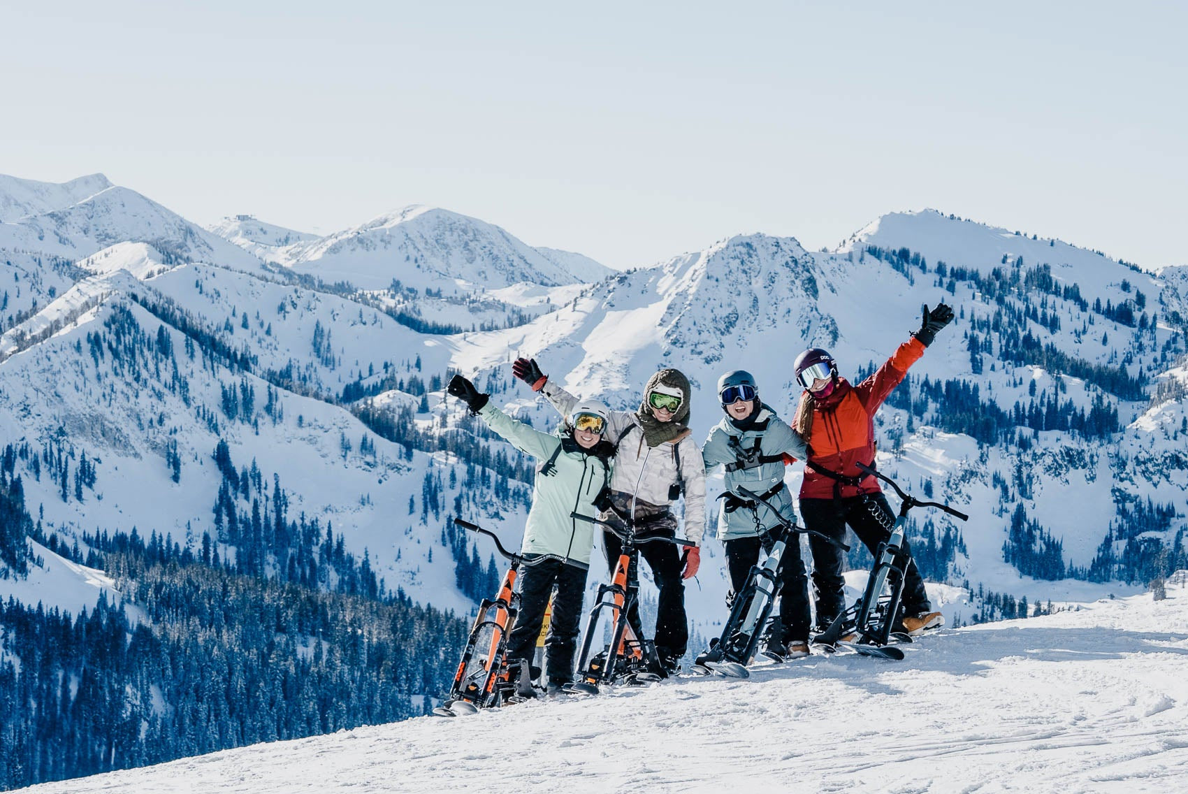 Four skibikers pose together on a snowy mountain slope with majestic snow-covered peaks in the background. They are wearing colorful winter gear, including an orange jacket, a light blue jacket, and a gray jacket, and have their snowboards and ski bikes ready.
