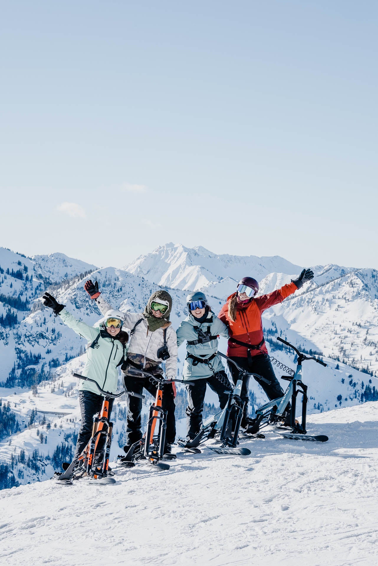 Four ski bikers pose together on a snowy mountain ridge with snow-covered peaks and a clear blue sky in the background. They are wearing winter sports gear and have their bikes beside them.
