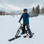 Person with a snow bike on a snowy slope with ski lifts and trees in the background
