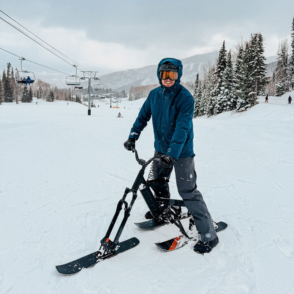Person with a snow bike on a snowy slope with ski lifts and trees in the background